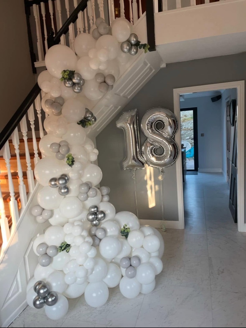 White and silver balloon garland flowing down a staircase beside giant silver 18 numbers