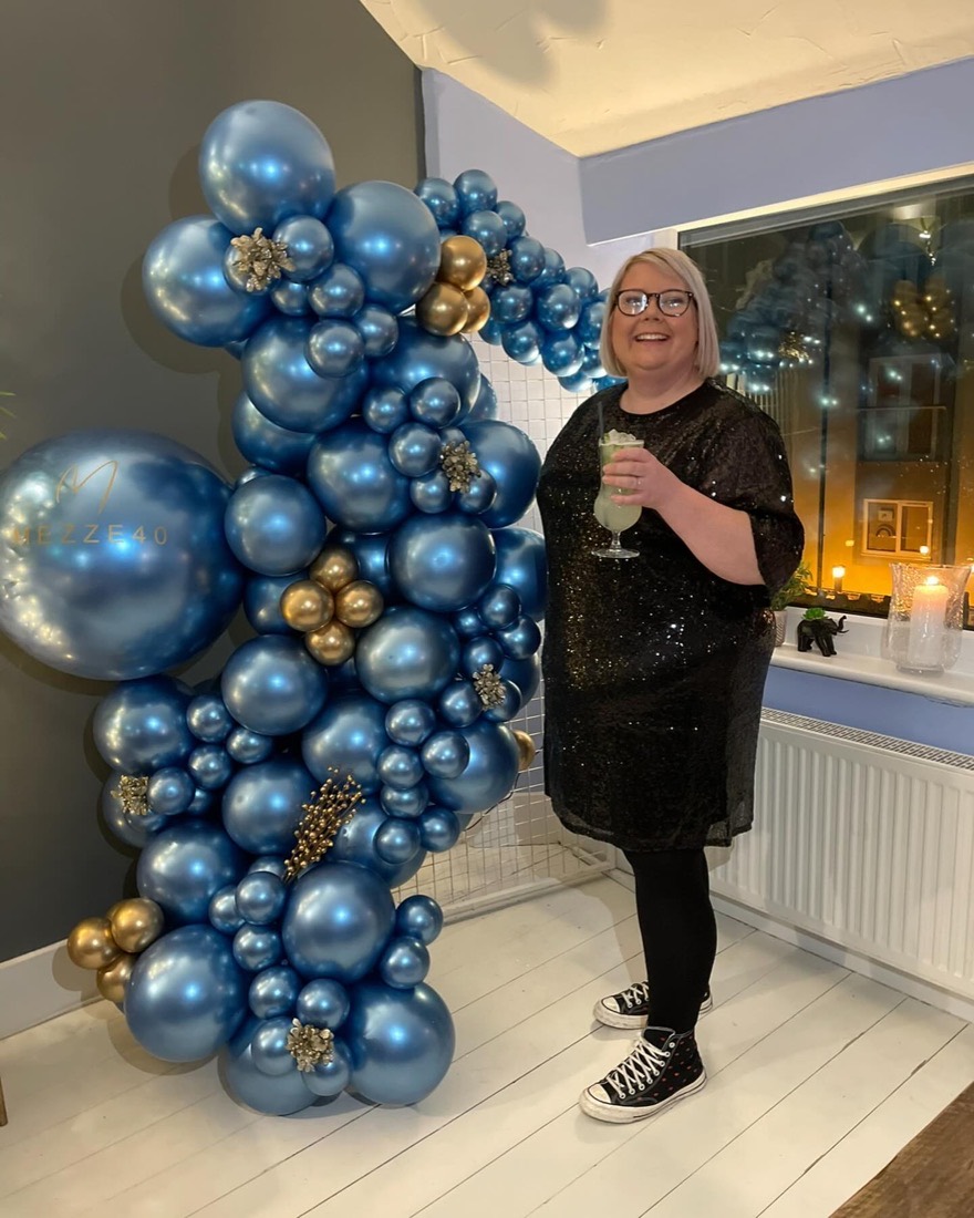 Amy beside a tall garland of chrome blue and gold balloons