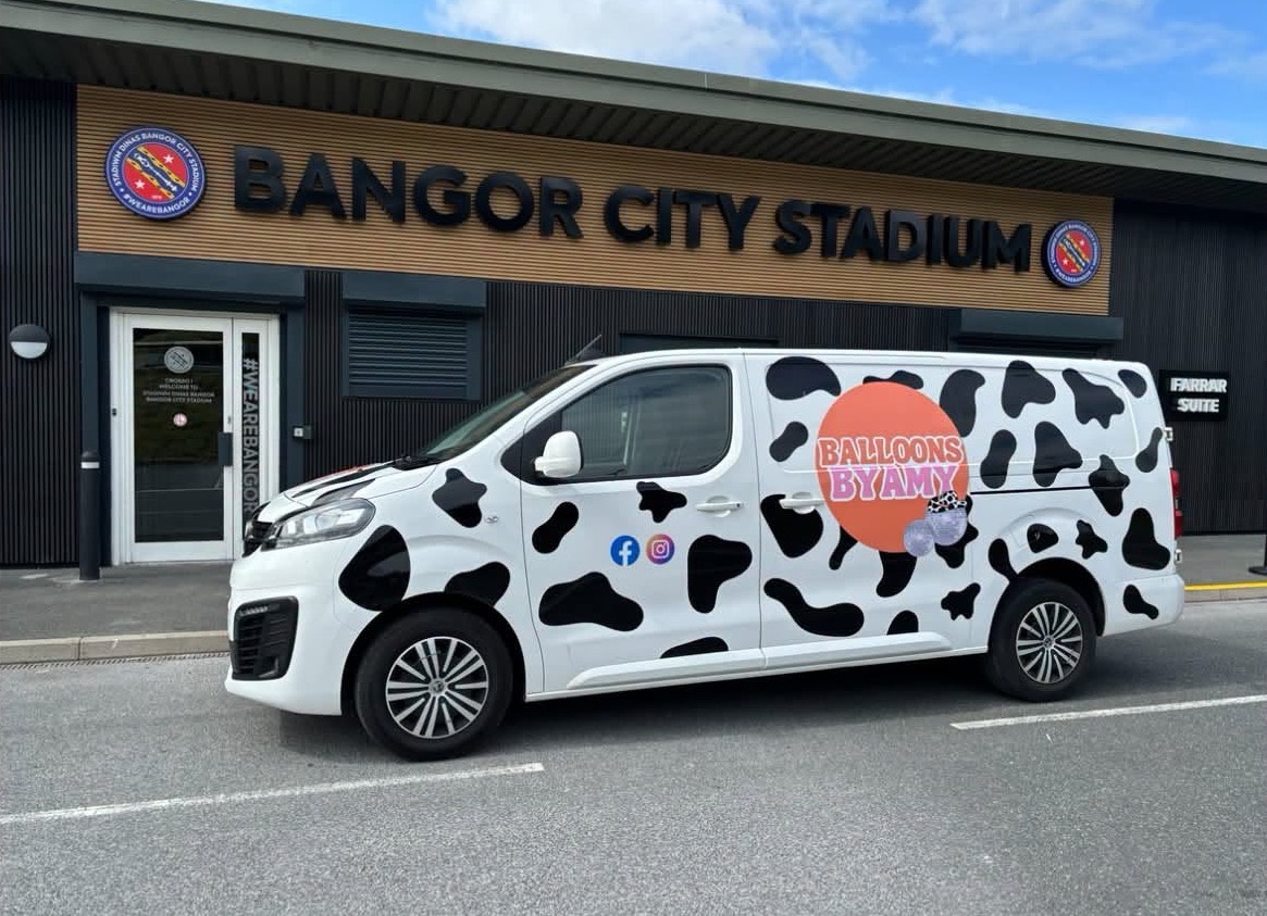 The Balloons By Amy van, white with big black cow patches and the orange logo, parked outside Bangor City Stadium